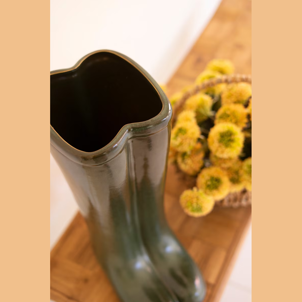 Glossy dark green ceramic rain boot vase on a wooden surface next to yellow flowers in a basket.