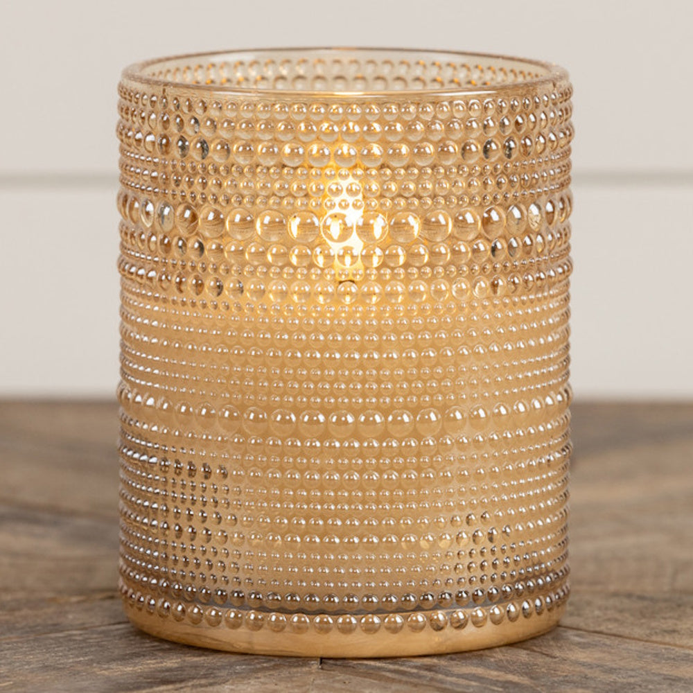 Lit candle in a champagne-tinted, beaded glass holder resting on a wooden surface.