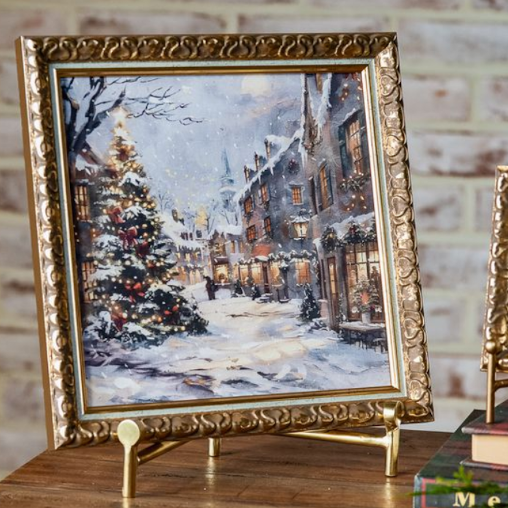 Framed art print depicting a snowy Christmas village scene, showcased in an ornate gold frame on a stand.