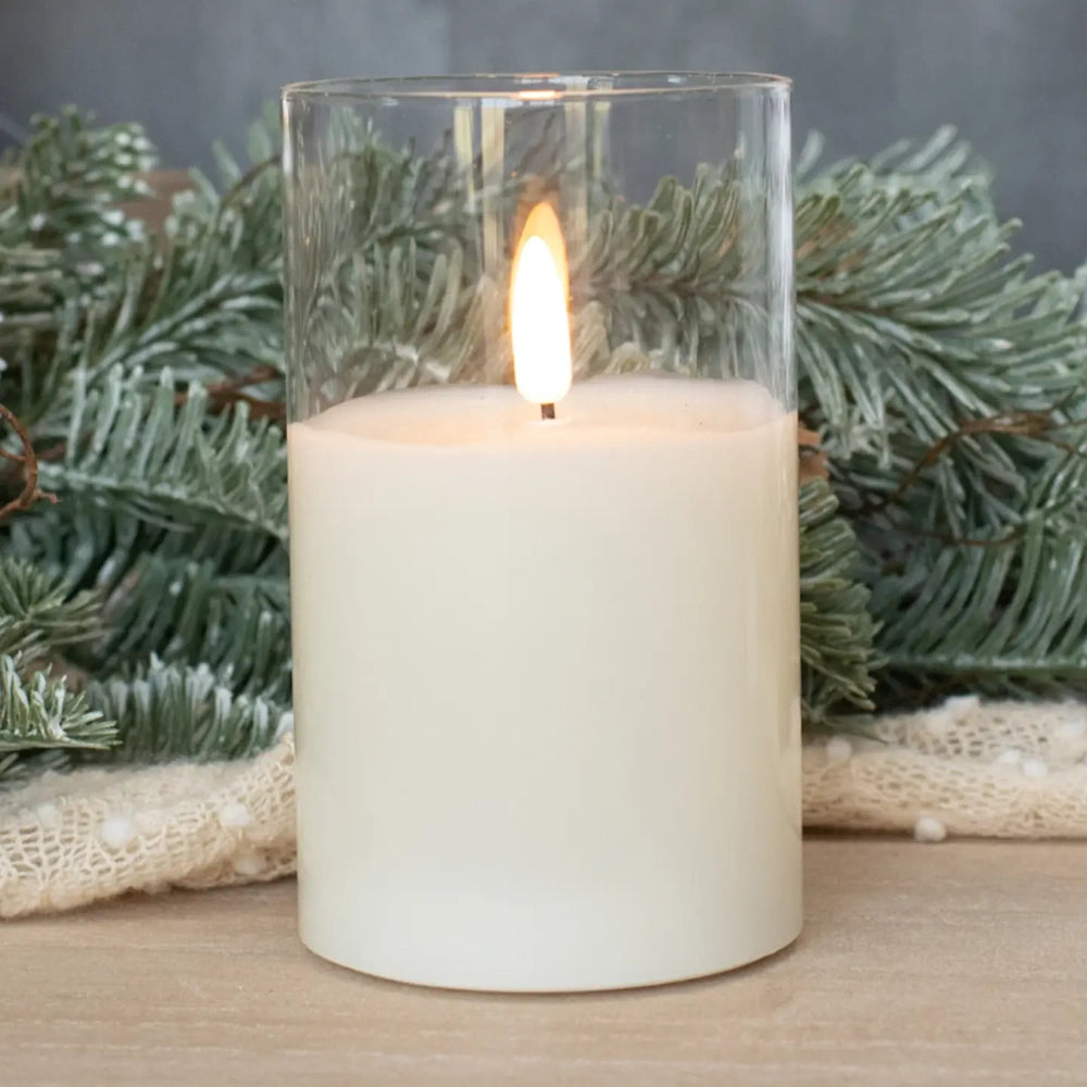 Lit white flameless pillar candle inside a clear glass cylinder in front of evergreen branches.