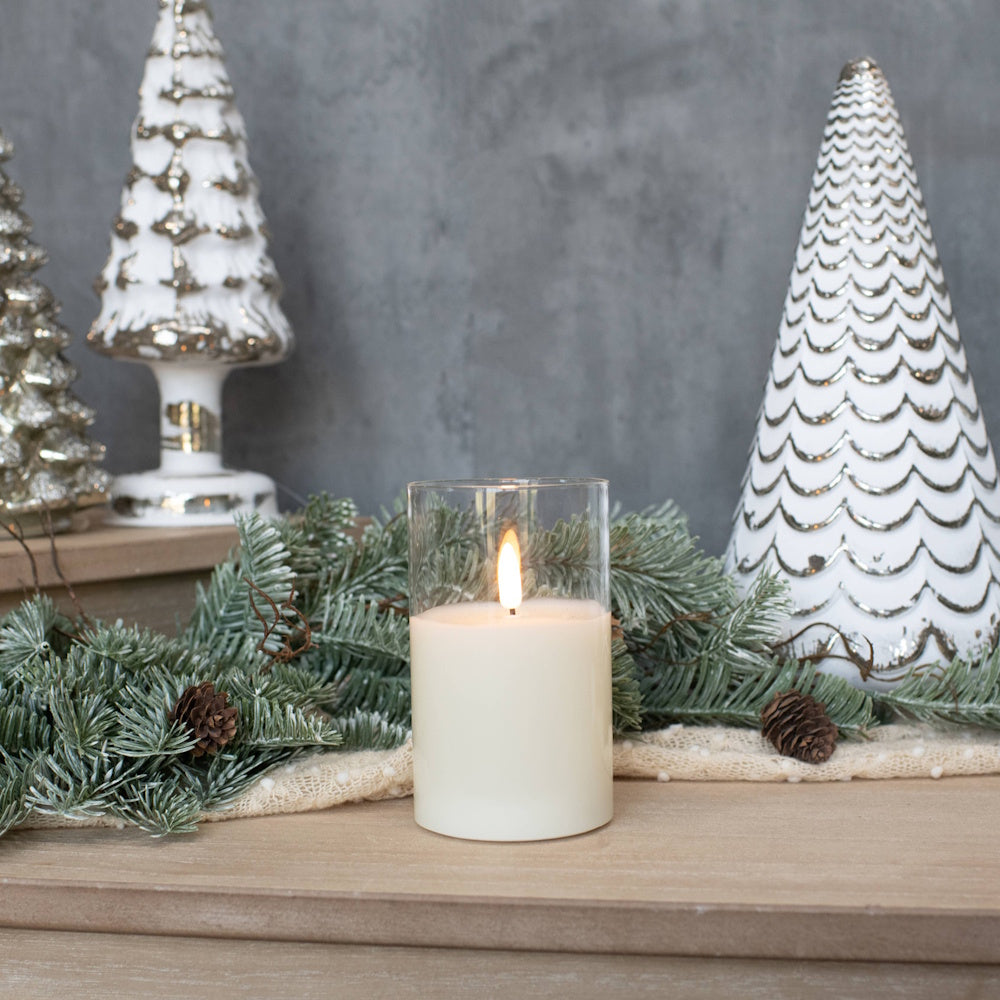 Lit pillar candle in a clear glass cylinder surrounded by evergreen pine garland and decorative Christmas trees.