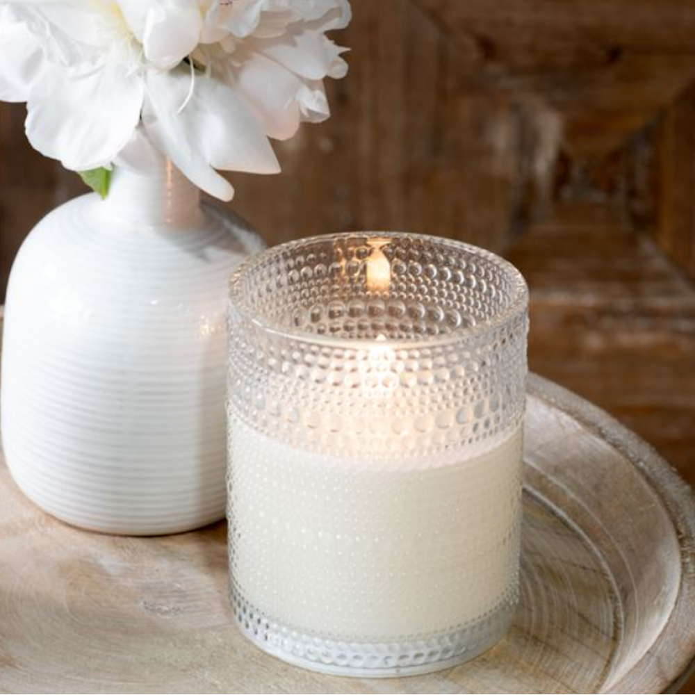 Tall lit LED candle in a clear hobnail beaded glass vessel sitting next to a white vase.