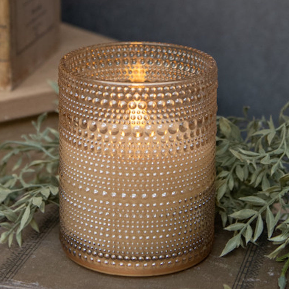 Lit candle glowing inside a champagne-gold beaded textured glass jar, framed by faux greenery.
