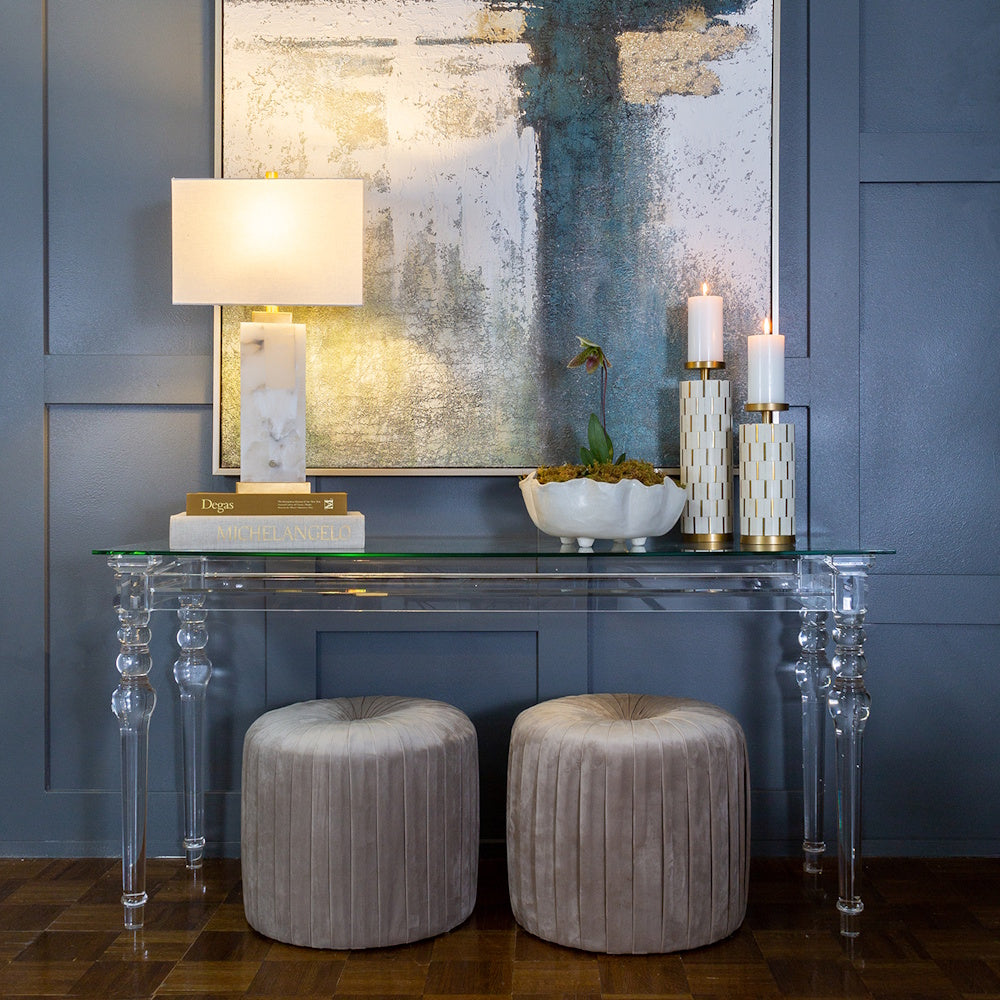 Modern acrylic console table styled with a white marble lamp, decorative bowls, and two velvet ottomans.