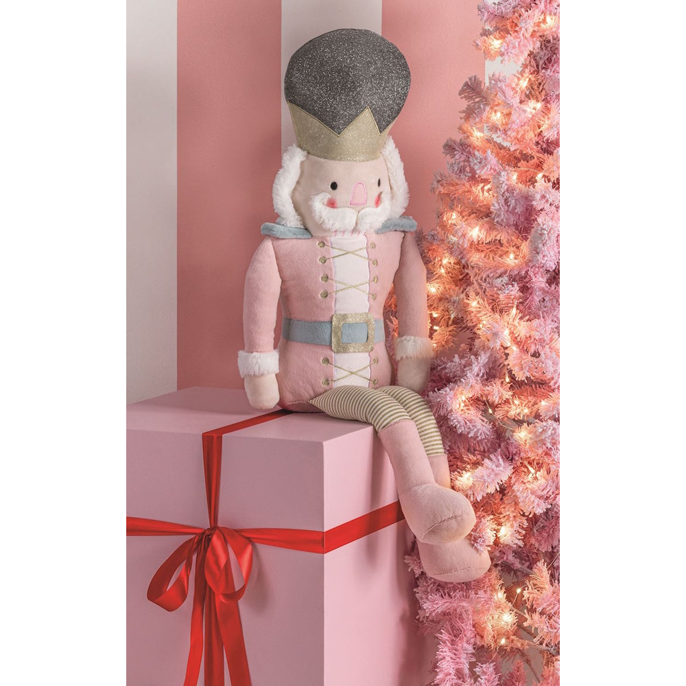 Pink nutcracker plush doll sitting on a large gift box next to a lit pink Christmas tree.