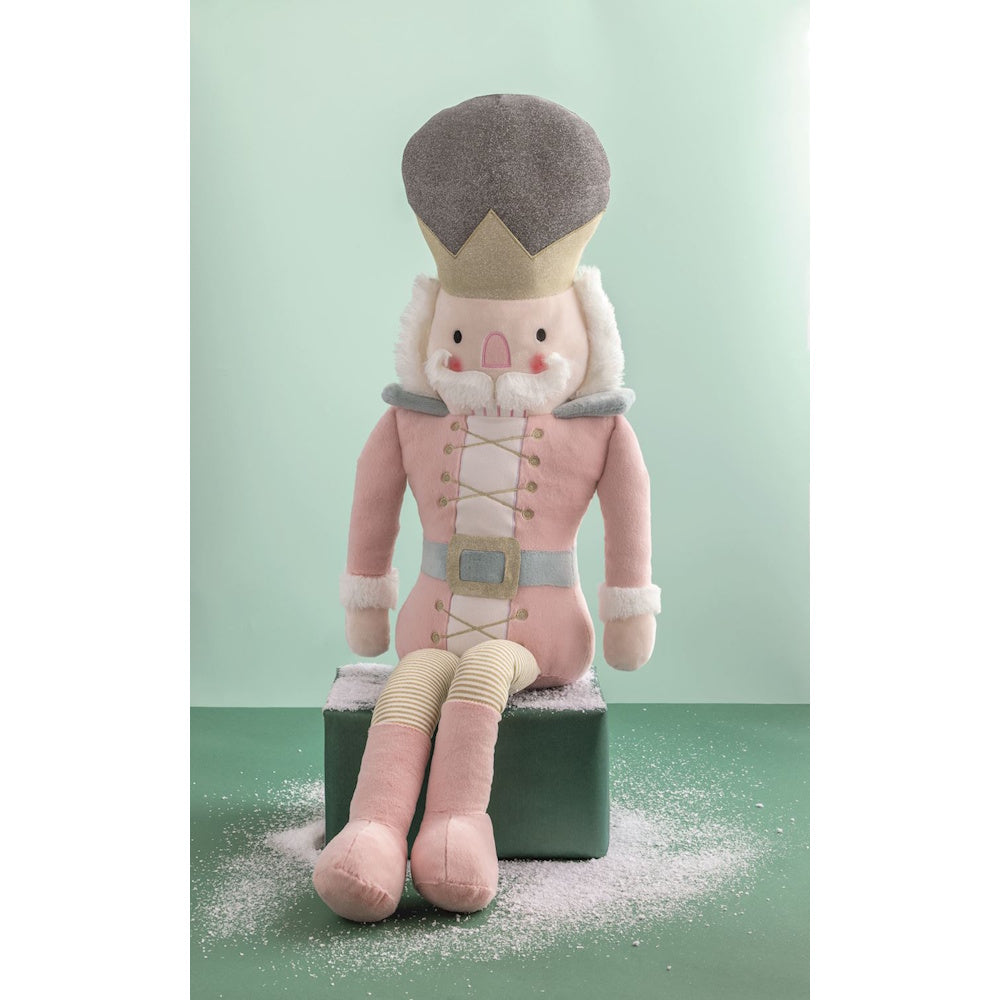Plush pink nutcracker doll in a gold crown sitting on a block with faux snow.