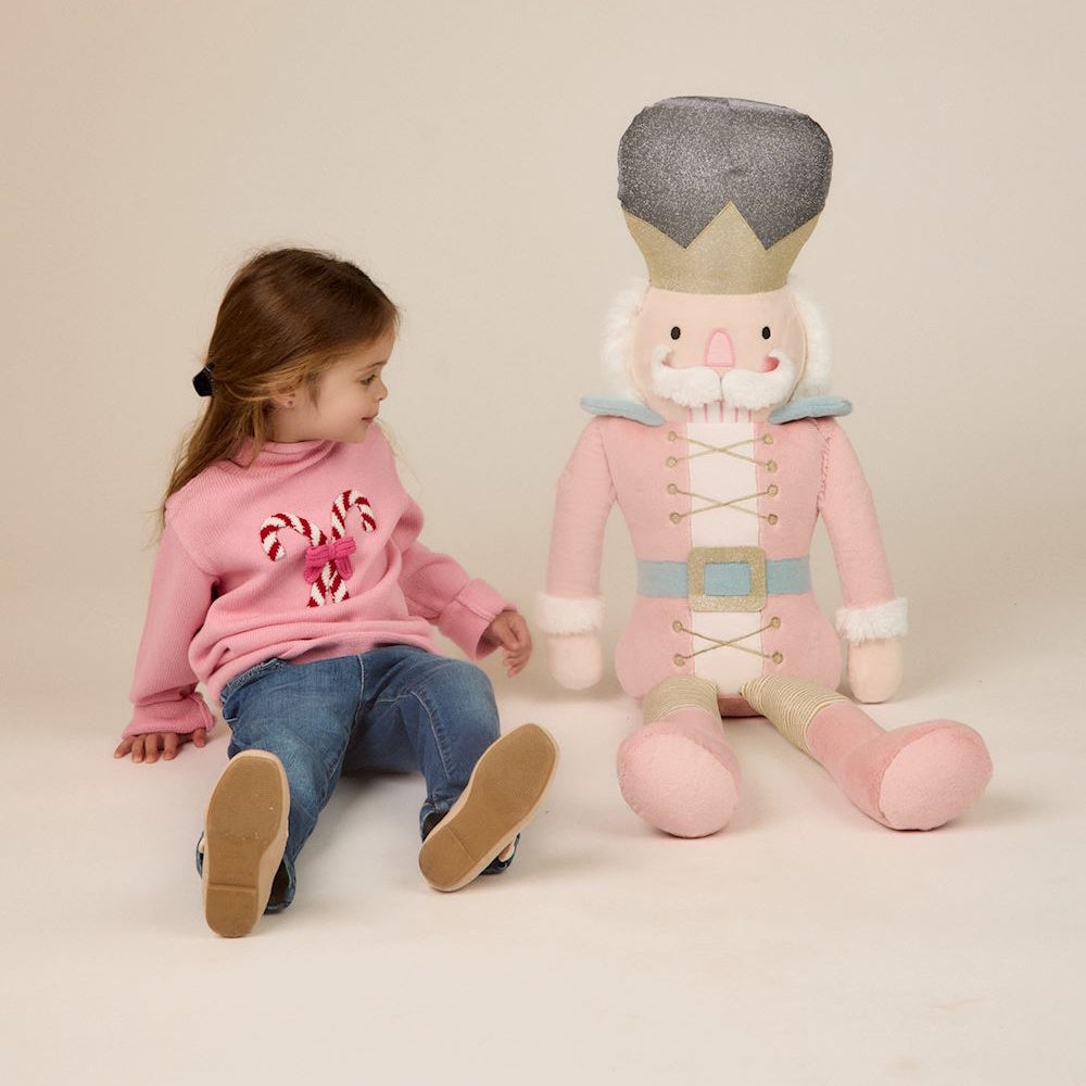 Young girl in a pink sweater sitting next to a large plush pink nutcracker doll.
