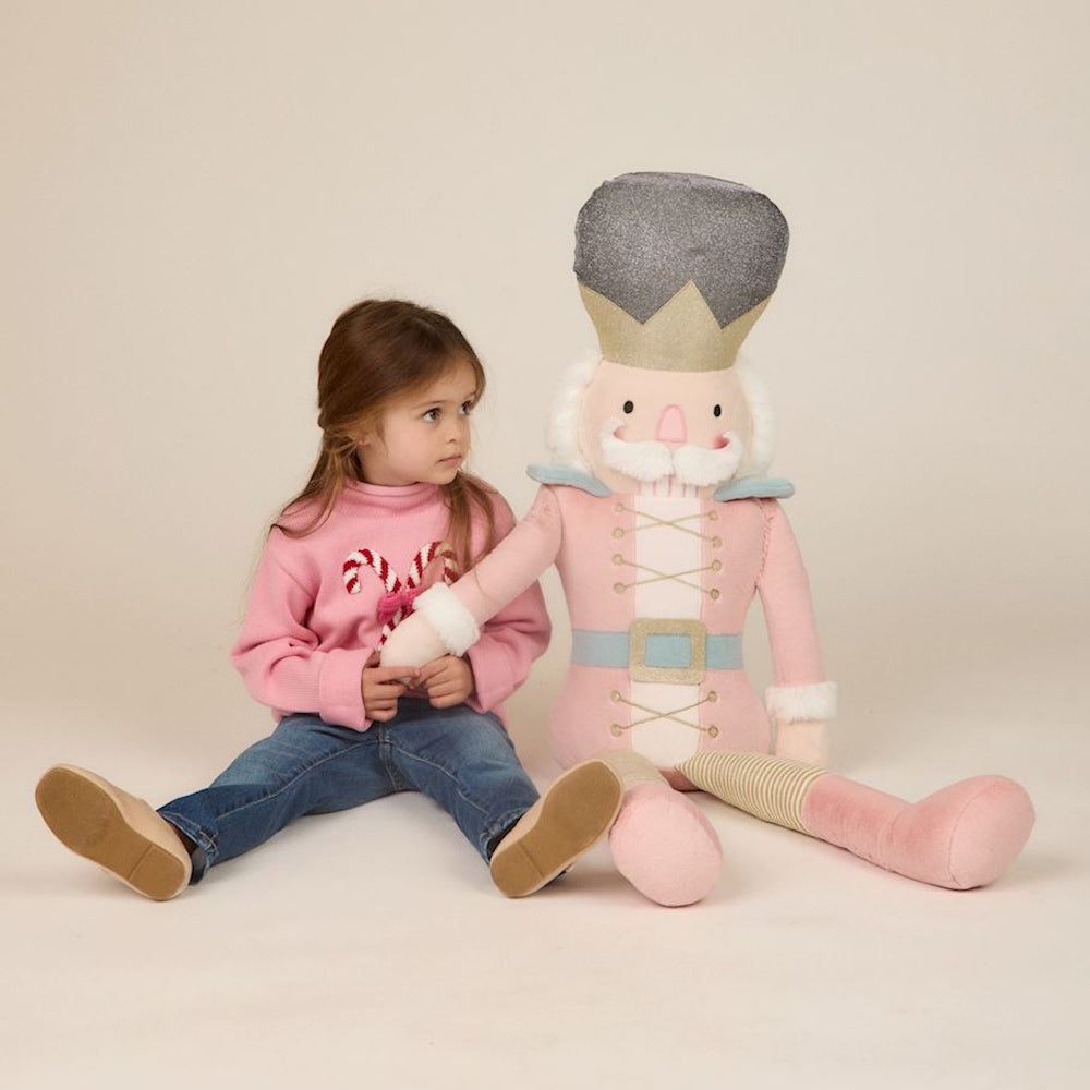 A young girl in a pink sweater sits beside a large plush pink nutcracker doll.