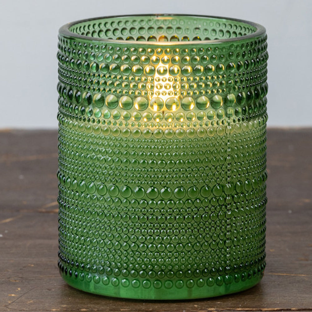 Lit candle inside a pine green beaded glass holder sitting on a dark wooden surface.