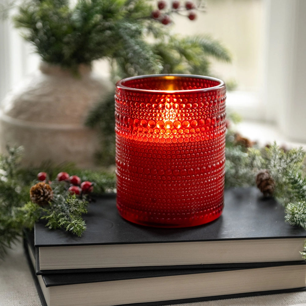 Glowing red dotted glass candle holder sitting on stacked books surrounded by festive evergreen greenery.