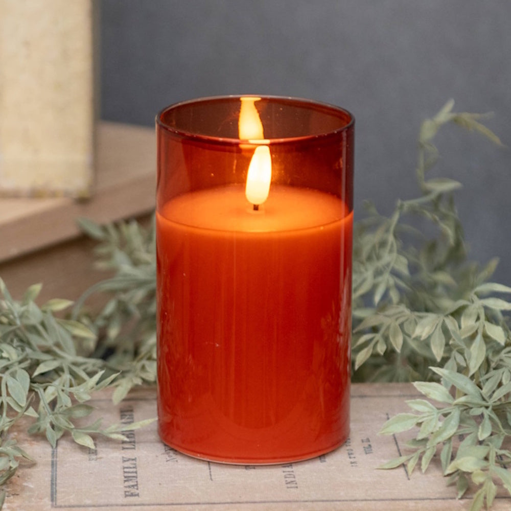 Lit orange candle inside a sienna glass holder surrounded by faux greenery on a vintage book.