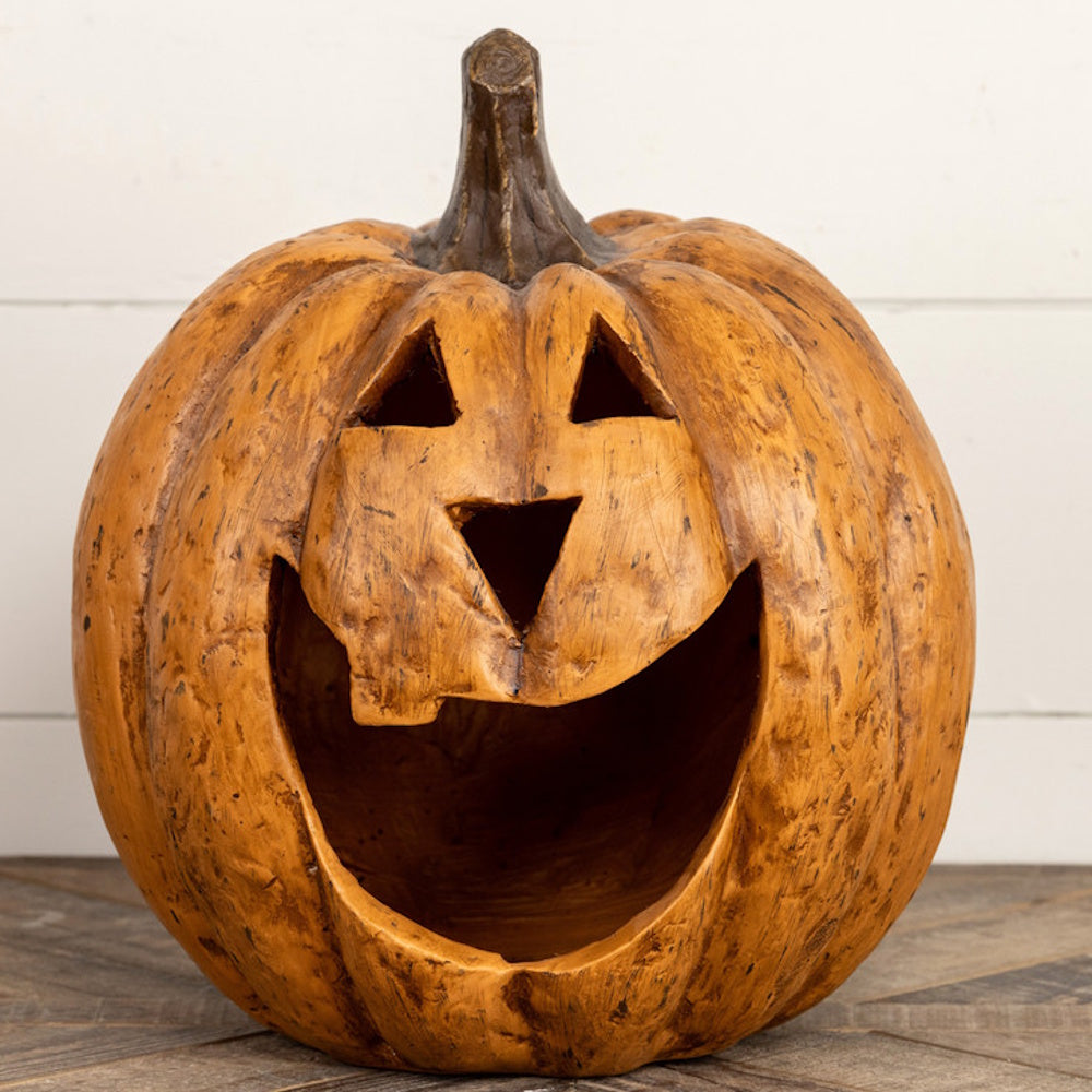 A carved rustic jack-o'-lantern with a wide smiling mouth sitting on a wooden surface against a white wall.