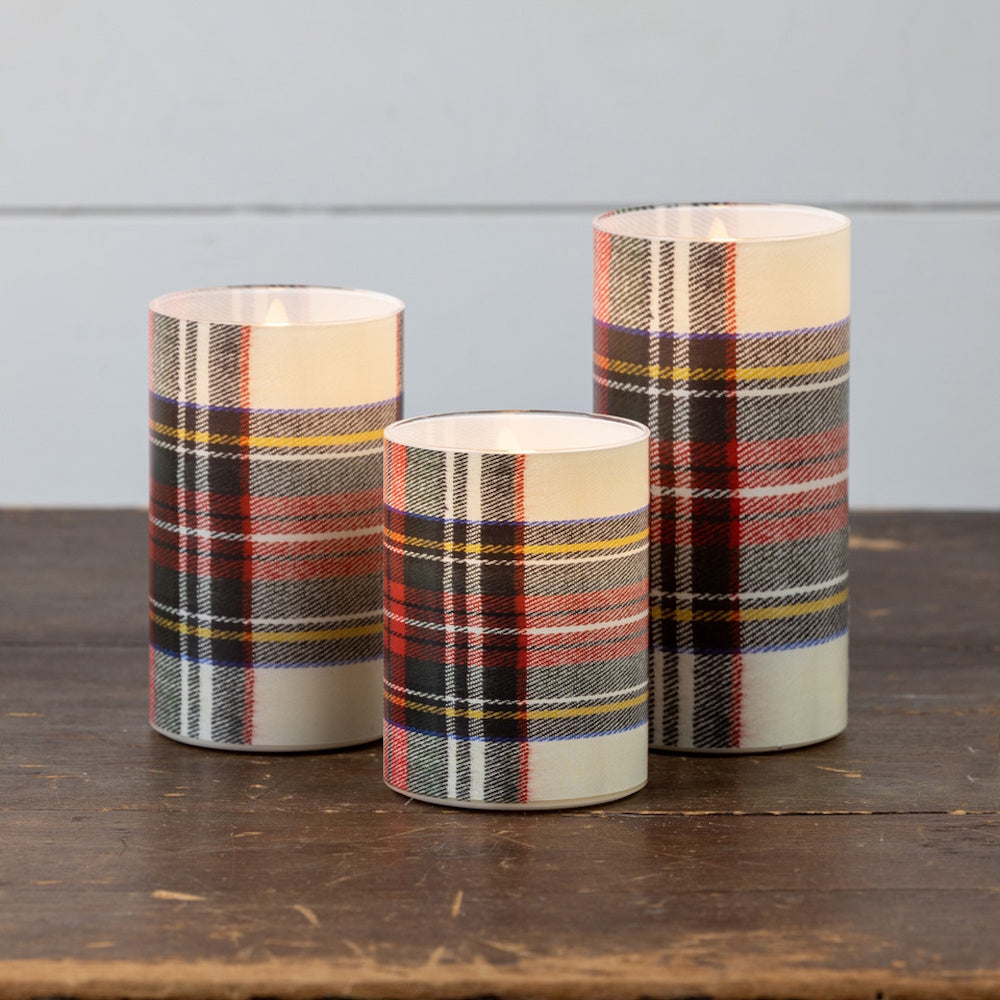 Three glowing white tartan plaid pillar candles of varying heights on a rustic wooden table.