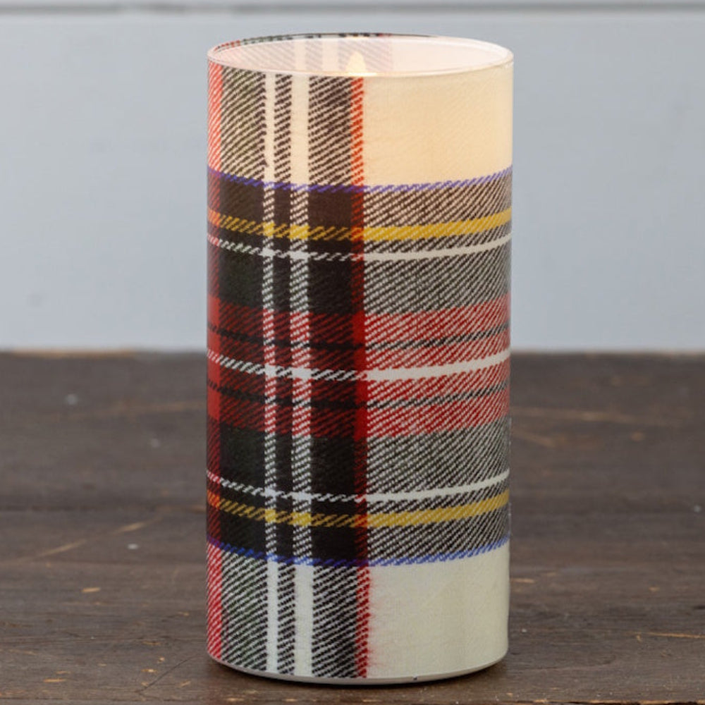 Lit white tartan plaid glass cylinder candle standing on a rustic wooden table.