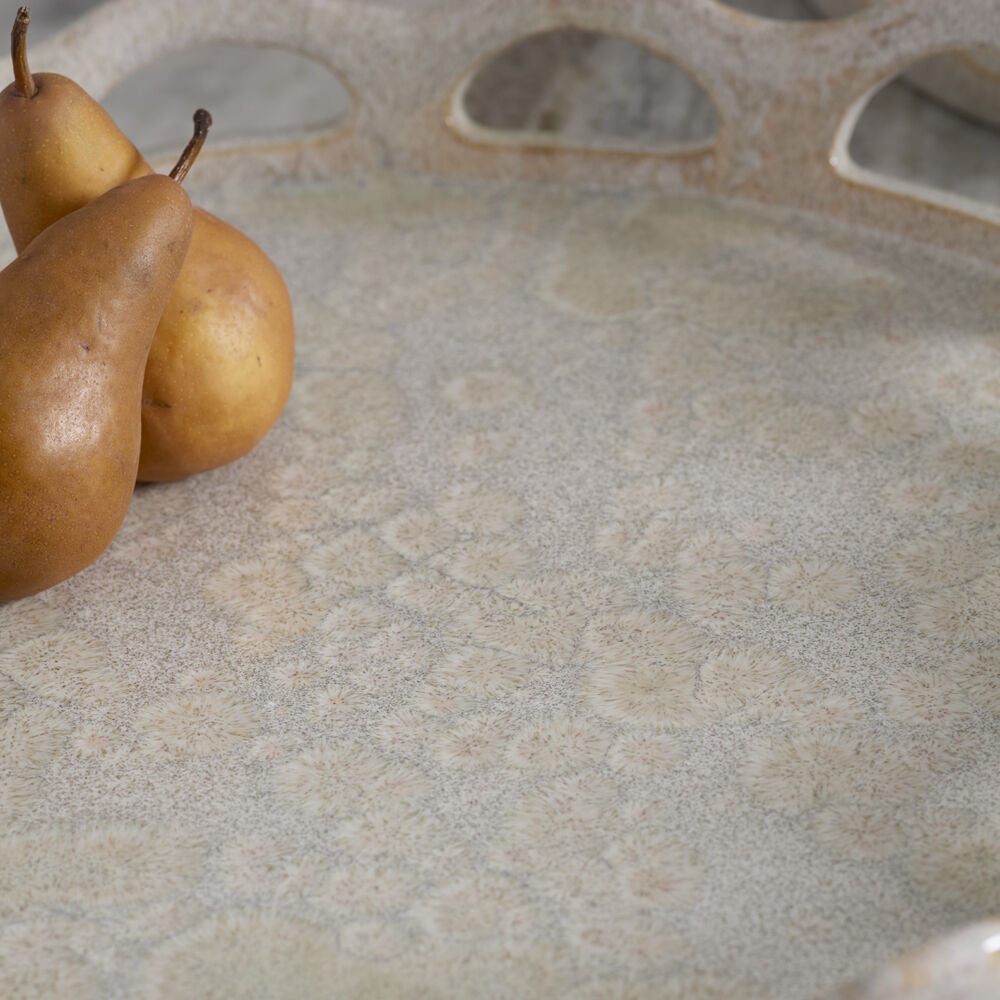 Close-up of a speckled, neutral-toned ceramic tray with decorative cutout handles and three golden-brown pears.