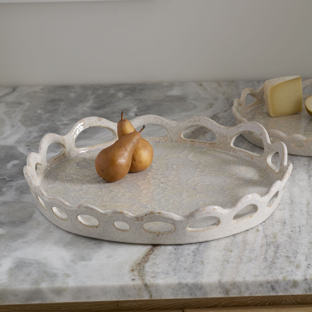 Round ceramic tray with a scalloped, cutout rim holding two brown pears on a marble countertop.
