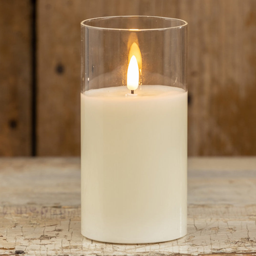 Large white pillar candle with a glowing flame inside a clear glass cylinder on a rustic wooden table.