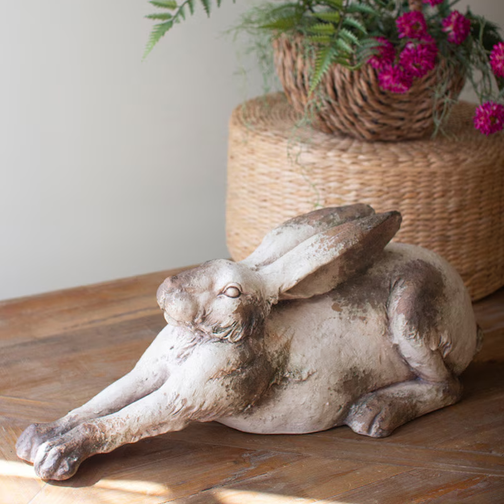 Stretching faux concrete rabbit statue on a wooden table with a wicker basket and pink flowers in the background.