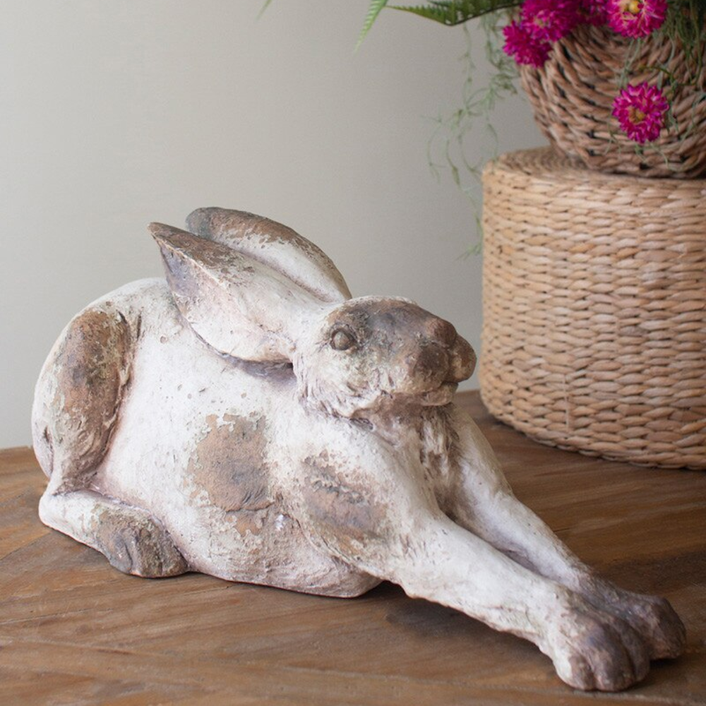Rustic faux concrete rabbit statue laying on a wooden surface next to a woven basket with pink flowers.