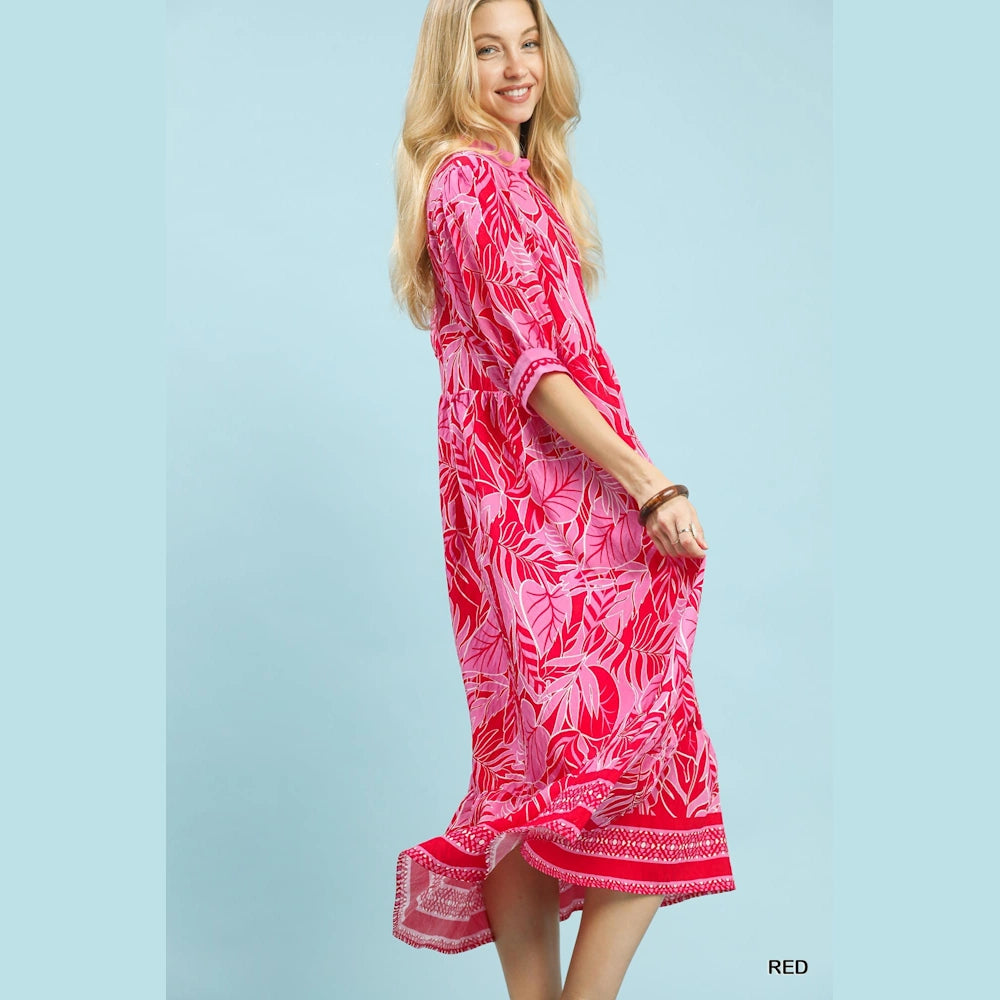 Woman wearing a pink and red leafy print v-neck midi dress with a decorative border hem against a light blue background.
