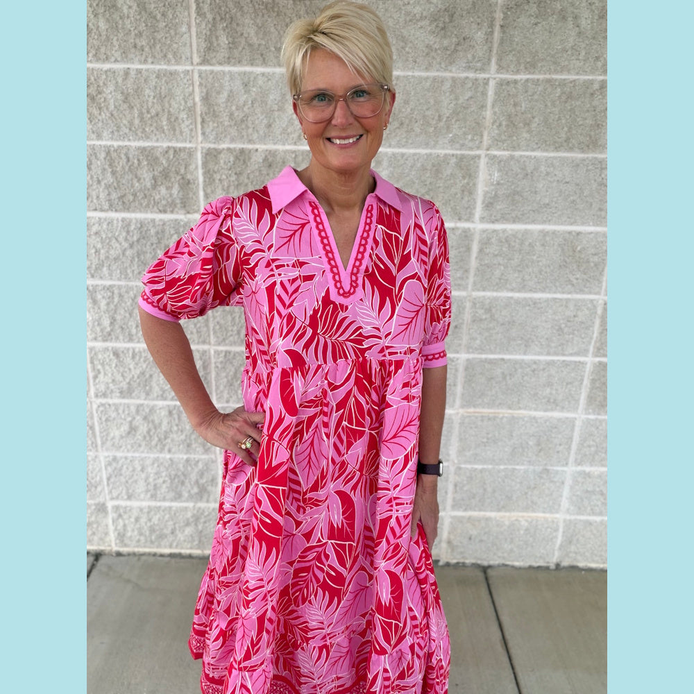 Blonde woman wearing a pink and red leafy print V-neck midi dress with puff sleeves and a collar against a brick wall.