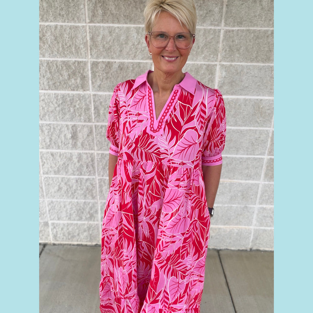 Woman in a pink leafy print V-neck midi dress with puff sleeves and a collar, standing against a white brick wall.
