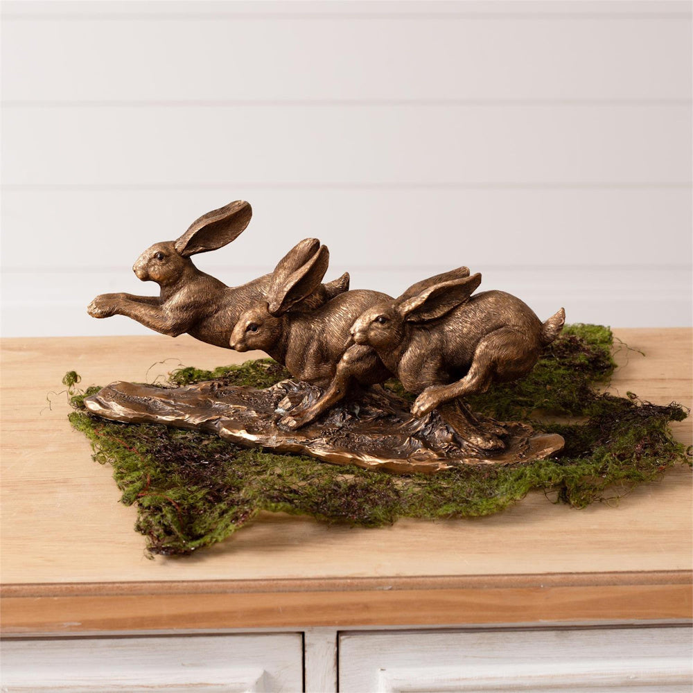 Bronze-tone leaping hares sculpture featuring three running rabbits on a mossy base for rustic shelf decor.