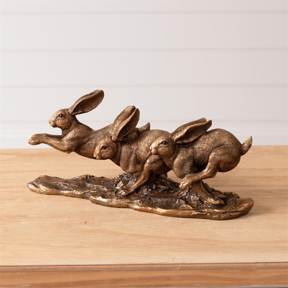 Bronze-tone sculpture of three leaping hares on a textured base, displayed on a wooden surface against a white wall.