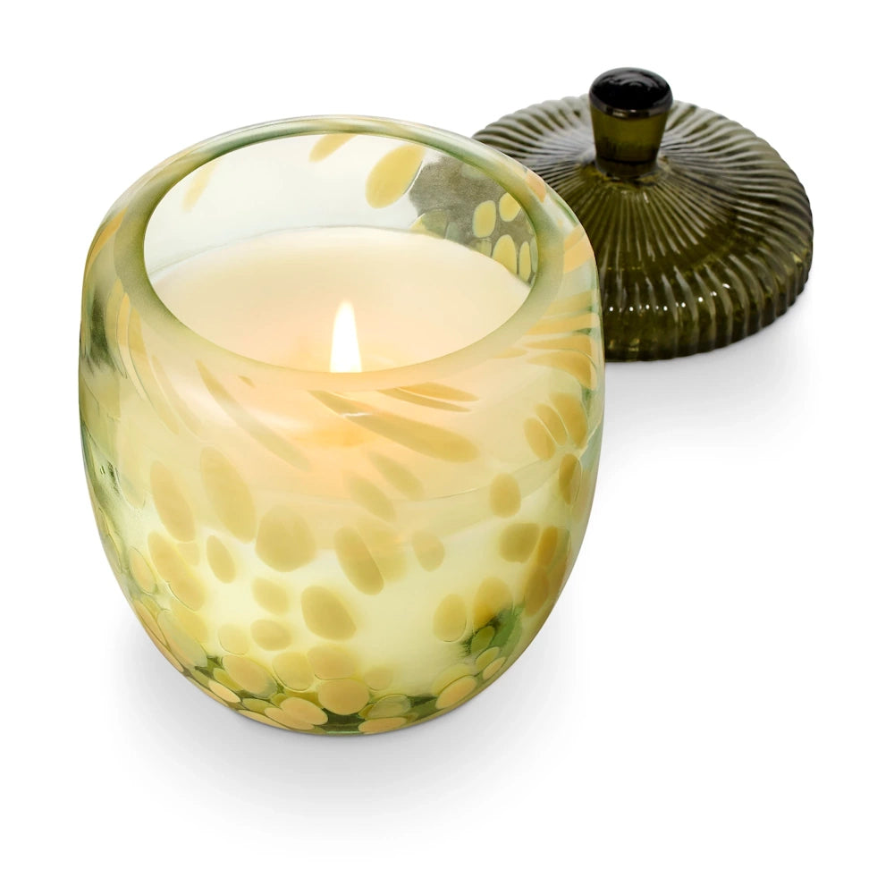 Lit candle in an acorn-shaped green speckled glass jar with a textured lid nearby.