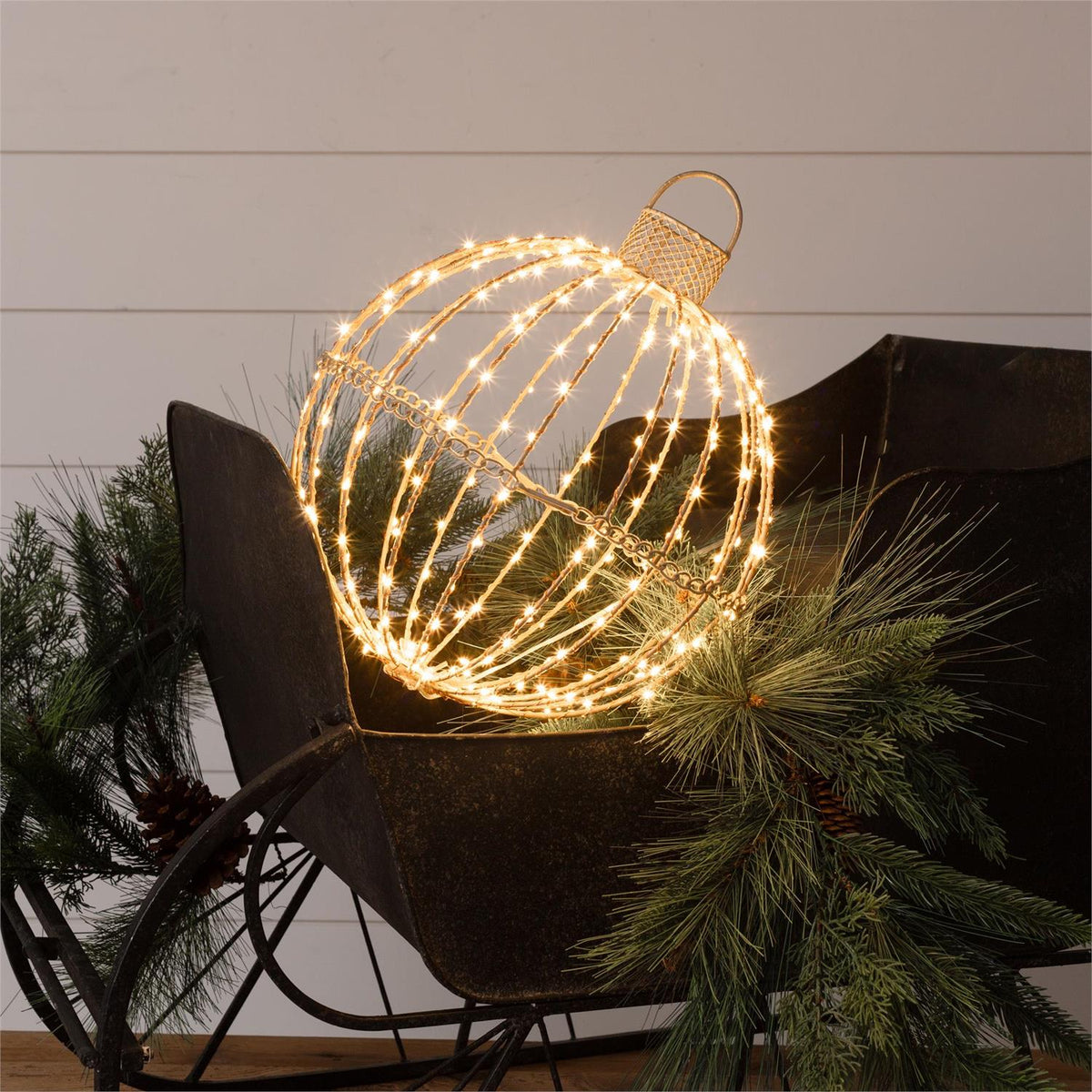 Illuminated warm-white LED wire sphere ornament resting in a vintage metal sleigh with pine greenery.