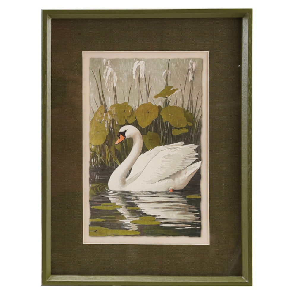 Framed artwork of a white swan swimming in a pond with lily pads and reeds, set against a dark mat.