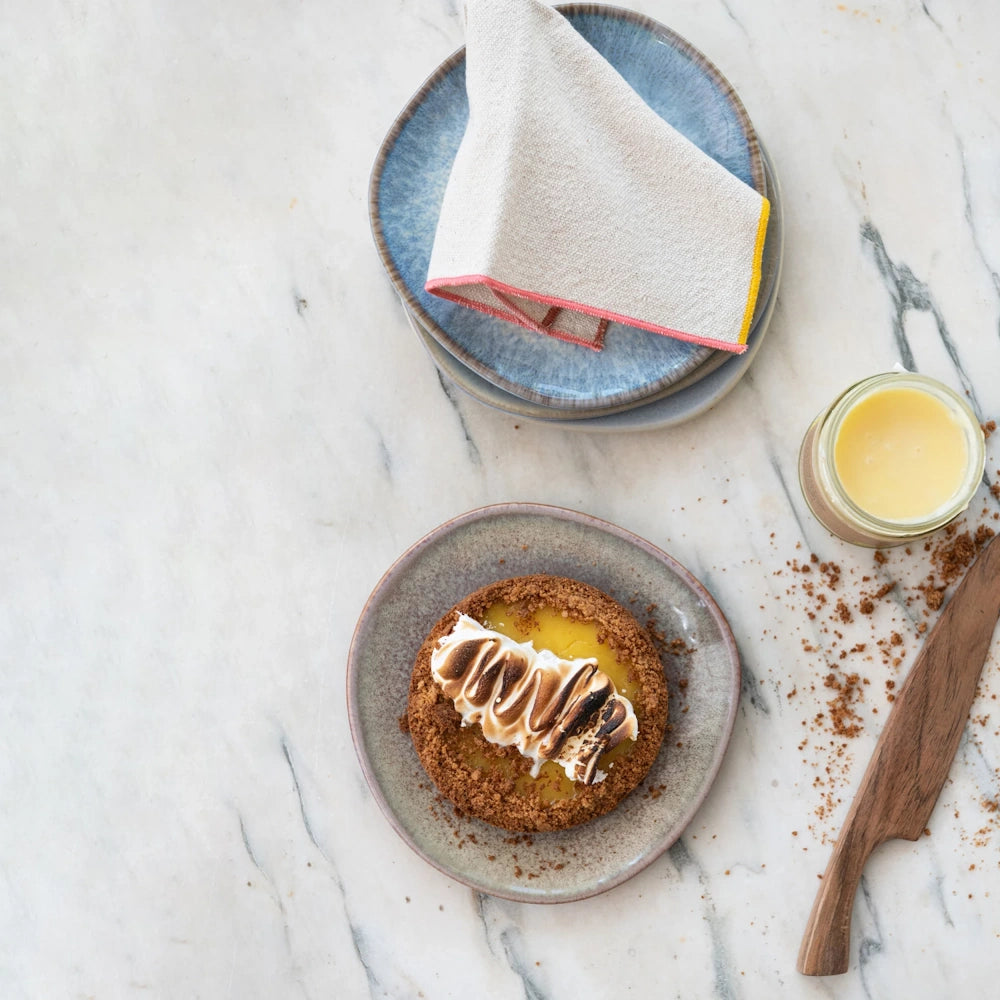 Lemon tart with toasted meringue on a grey plate next to a jar of lemon curd and a wooden knife on marble.