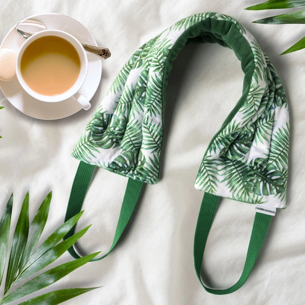 Lemon Lavender palm leaf print heated neck wrap with green handles next to a cup of tea on a white fabric surface.