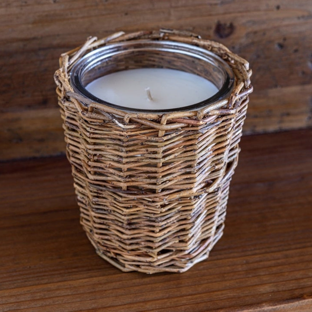 White wax candle inside a clear glass jar wrapped in a woven wicker sleeve on a wooden surface.
