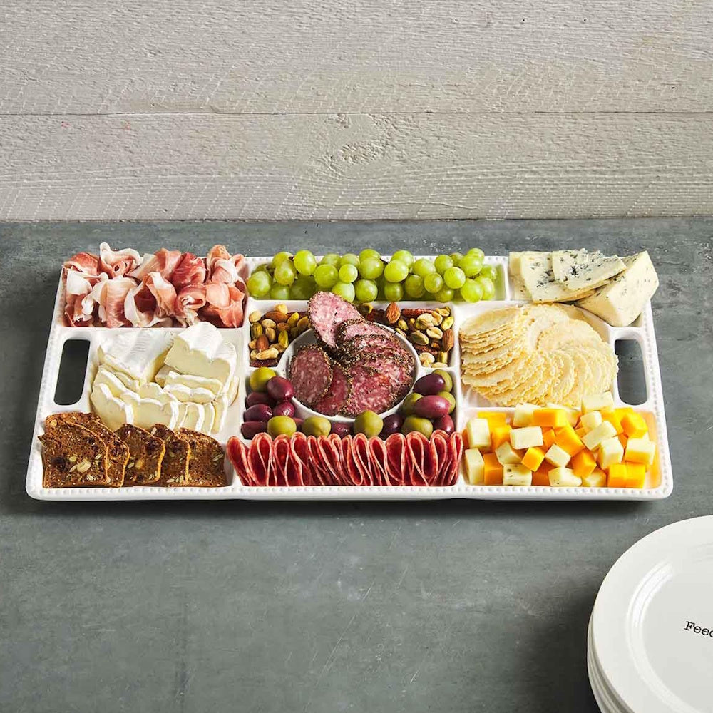White ceramic sectional charcuterie board filled with prosciutto, cheeses, grapes, salami, crackers, and olives.