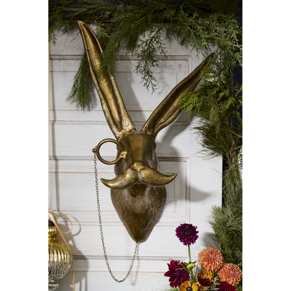 Gold hare wall mount with a monocle and mustache, displayed on a paneled door surrounded by greenery.
