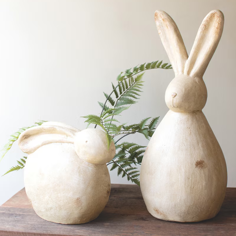 Pair of rustic, cream-colored terracotta bunny figurines and a green fern frond on a dark wood surface.