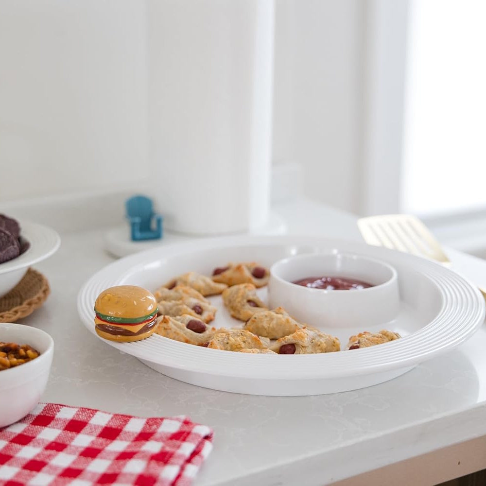 White serving platter with pigs in a blanket and ketchup, featuring a decorative ceramic burger charm on the rim.