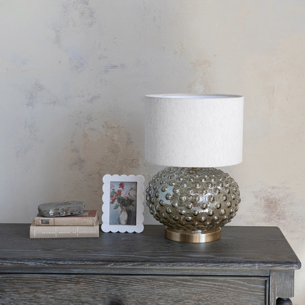 Smoky glass hobnail lamp on a dark wood dresser next to books and a framed picture against a textured wall.