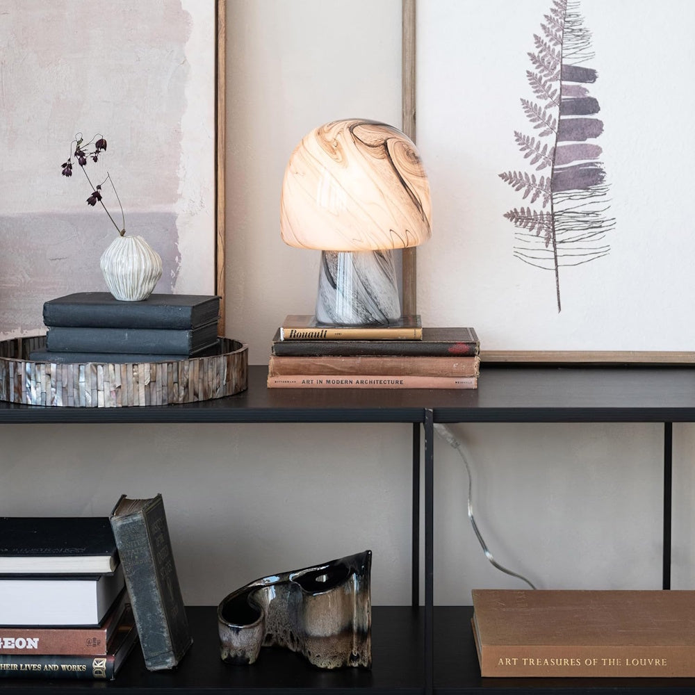 Hand-blown mushroom glass table lamp with brown swirls on a black shelf with art books and framed prints.