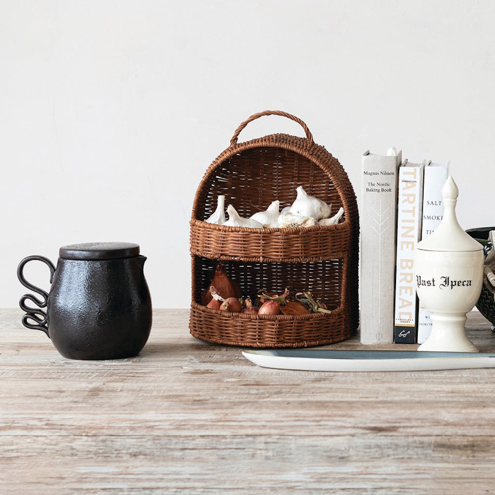 Hand-woven rattan 2-tier basket with garlic and shallots on a rustic wooden table with kitchen decor and books.