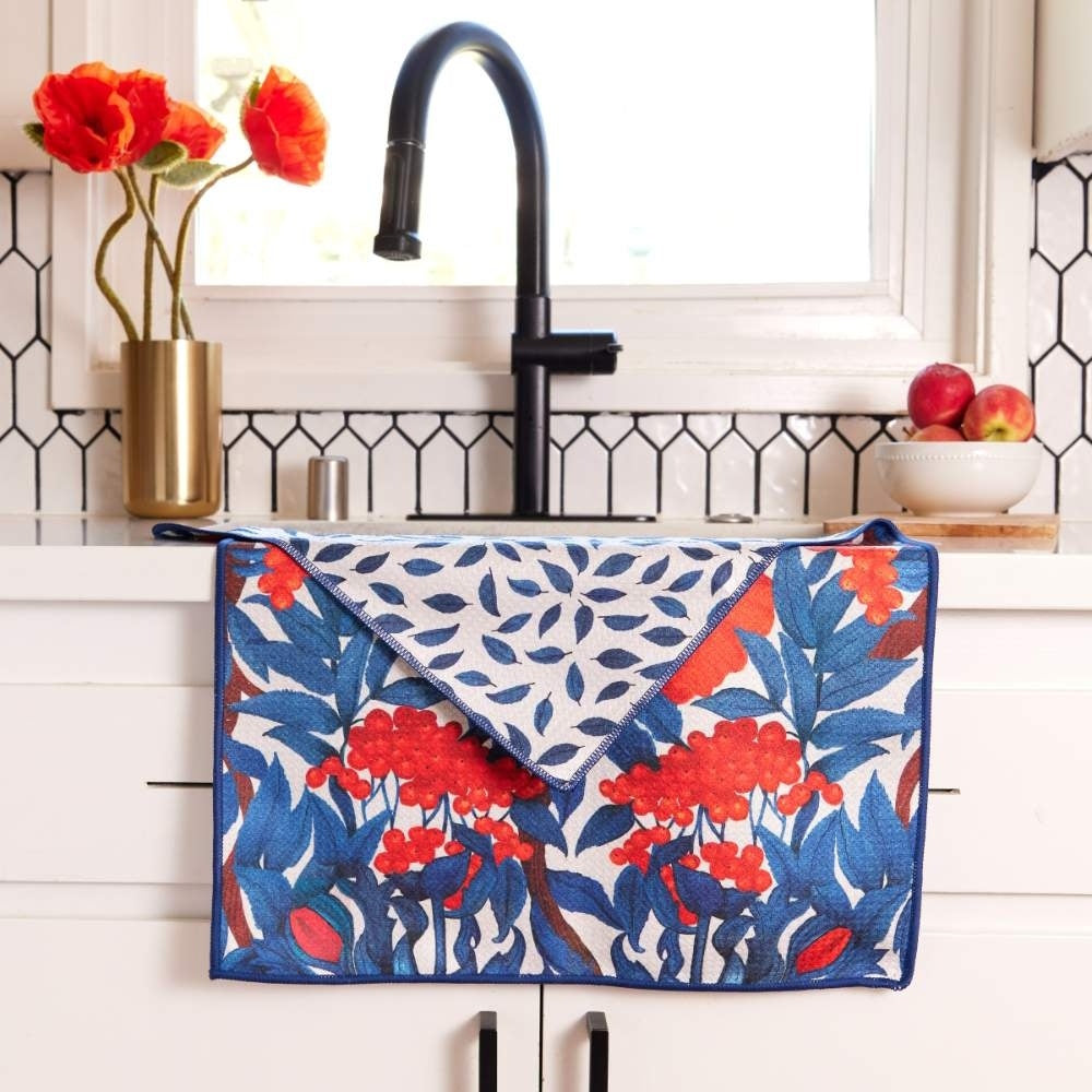 Icelandic poppies tea towel with blue leaves and red flowers draped over a white kitchen counter by a black faucet.