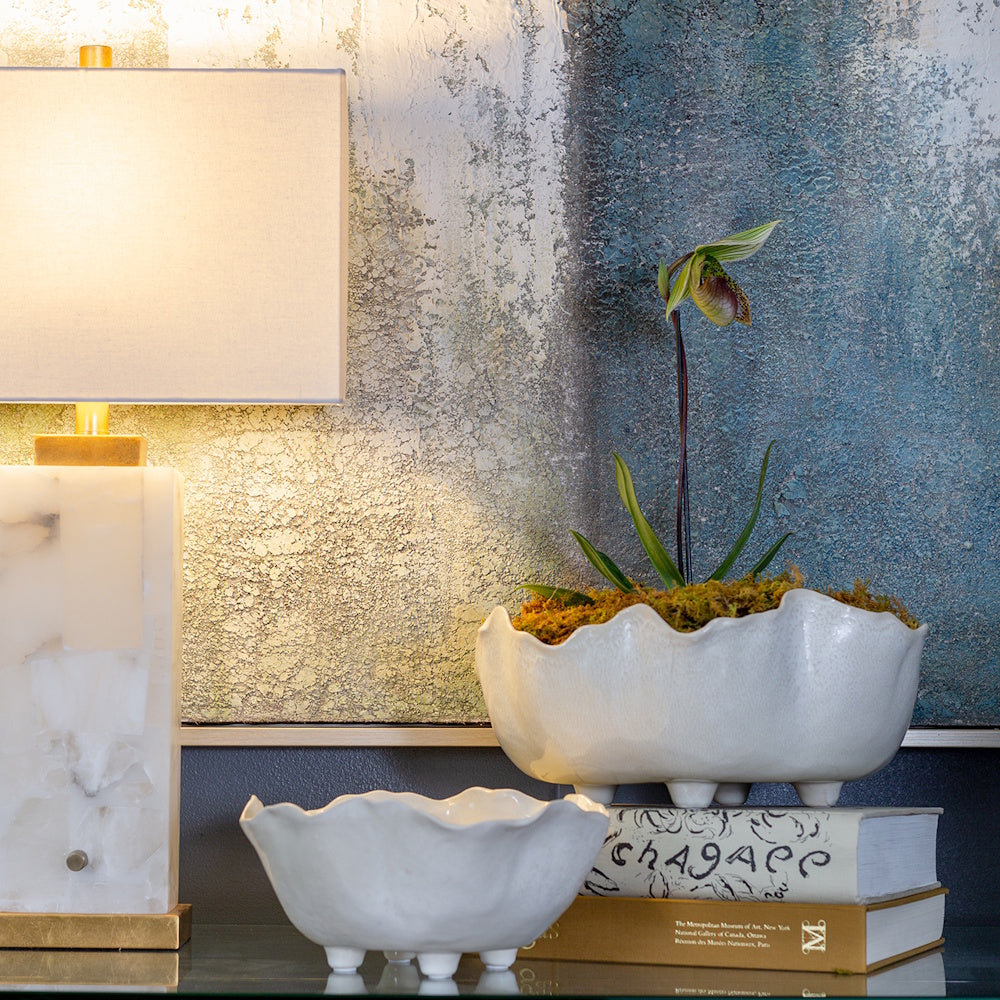 Two white ceramic Magnus bowls with scalloped edges used as a planter and decorative accent on books next to a lamp.
