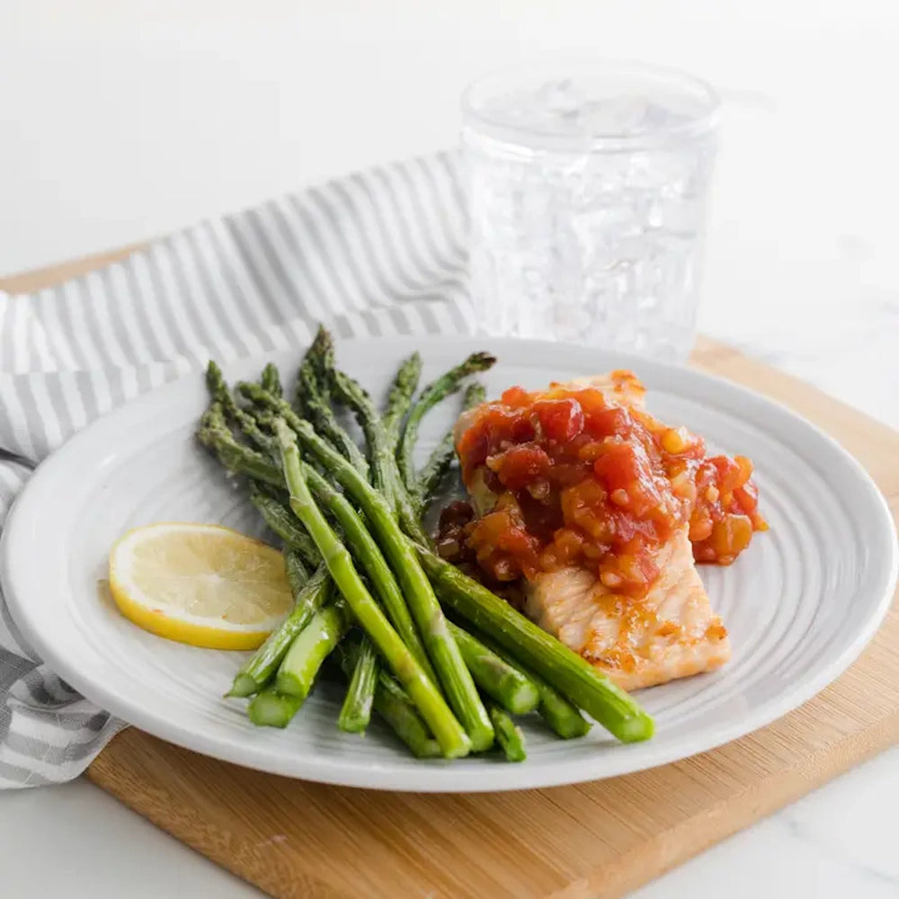 A white plate filled with cooked asparagus, a lemon slice, and a piece of salmon topped with fresh salsa.