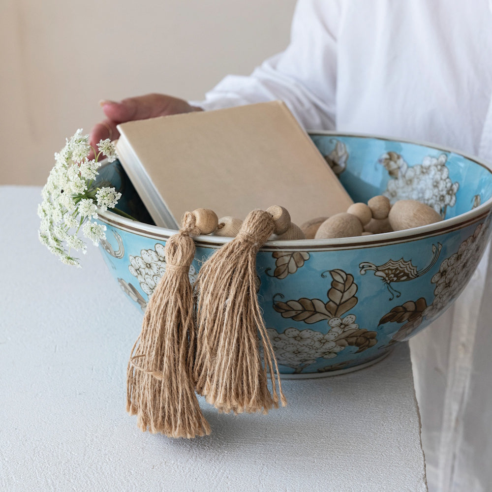 Blue floral stoneware bowl with cherry blossom and butterfly patterns, holding a book and decorative jute tassels.
