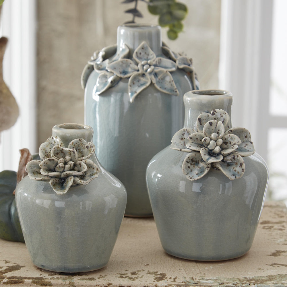 Three light blue ceramic bud vases with 3D floral accents and a crackle glaze finish on a rustic surface.