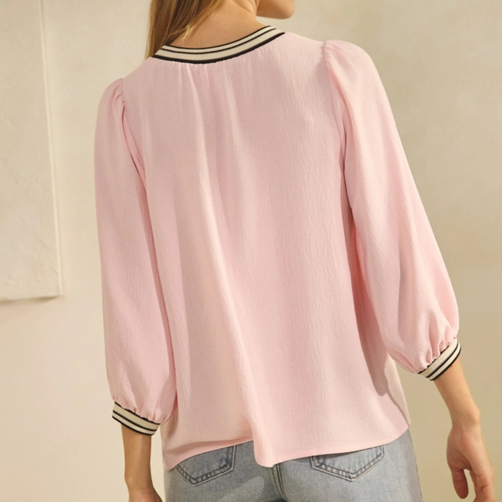 Back view of a woman wearing a light blush pink blouse with striped cuffs and neckline paired with denim jeans.