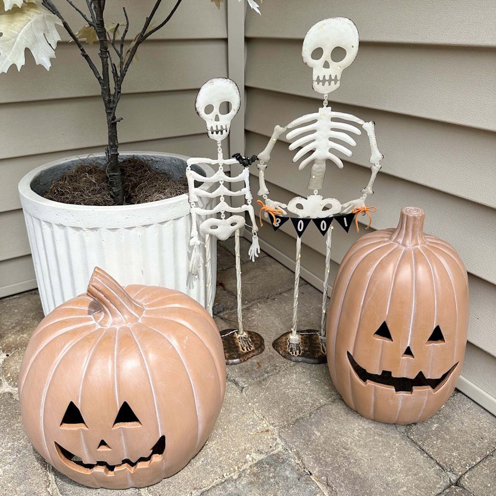 Two standing Halloween metal skeletons next to ceramic terracotta jack-o'-lantern pumpkins on a patio.