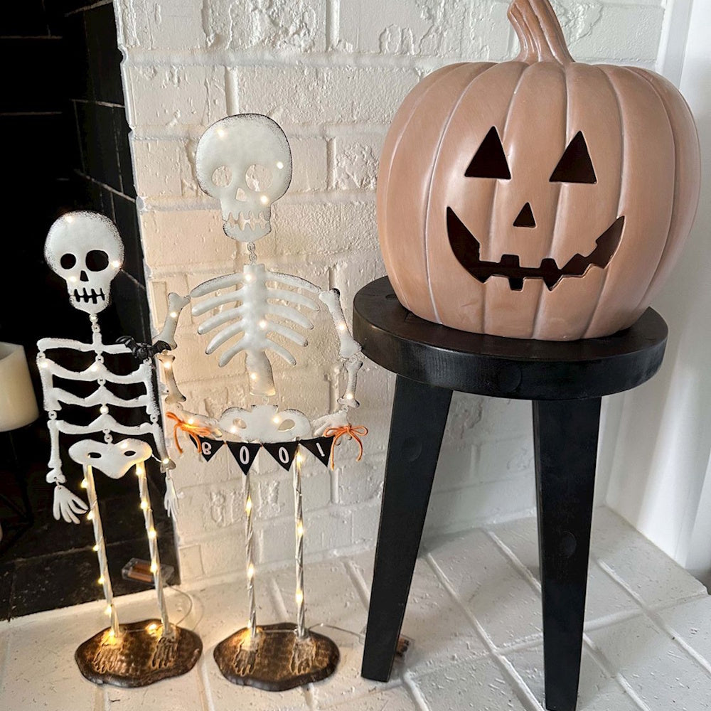 Light-up skeleton figurines next to a large jack-o'-lantern pumpkin on a black stool.