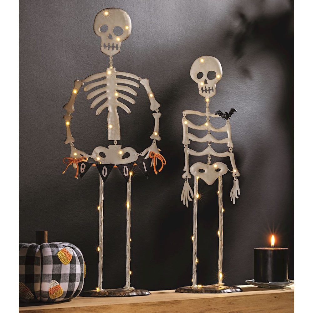 Two lit metal skeleton Halloween decorations on a shelf next to a plaid pumpkin and candle.